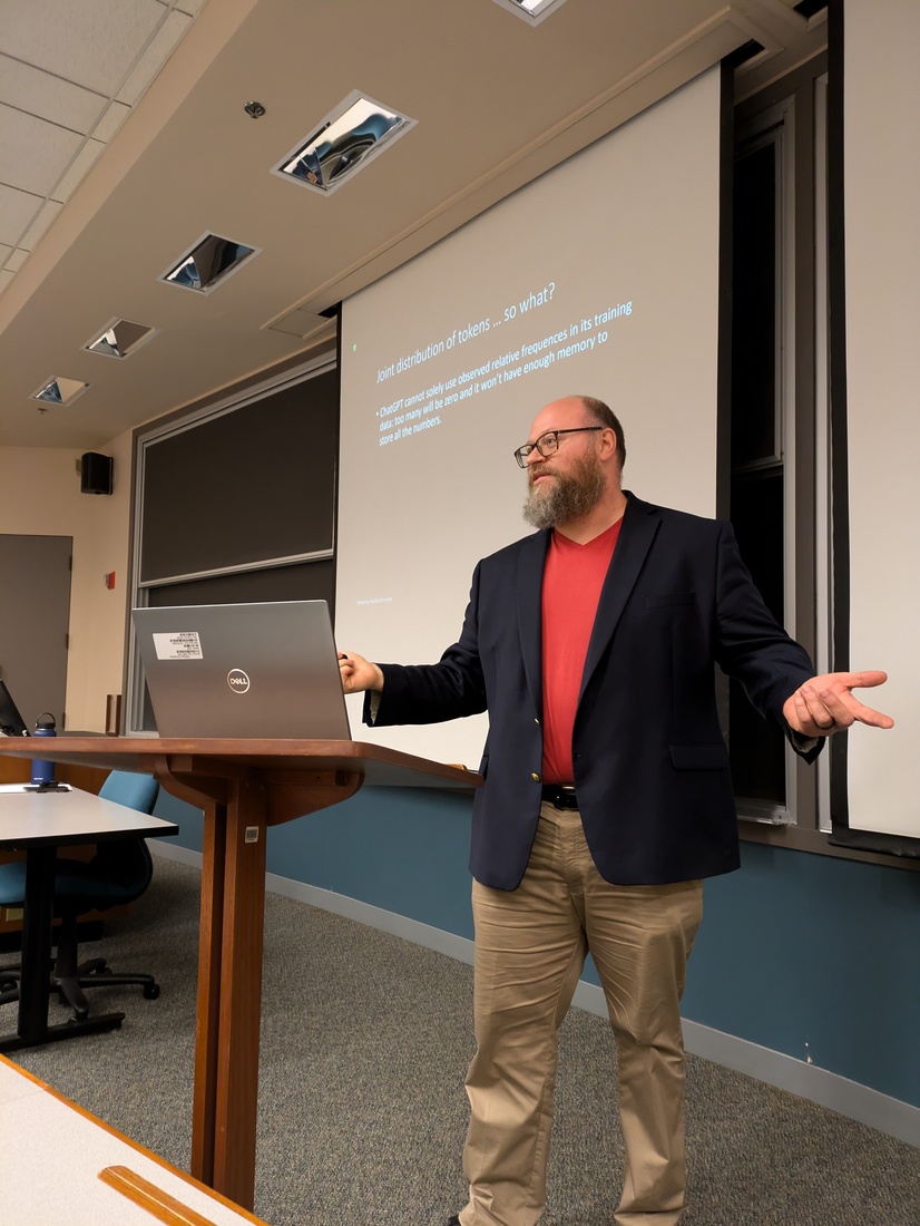 a man in a blazer and khakis lectures at the front of a classroom