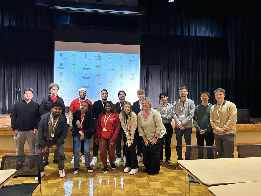 A group of the students who attended the NIU hackathon pose in front of a stage in a small auditorium