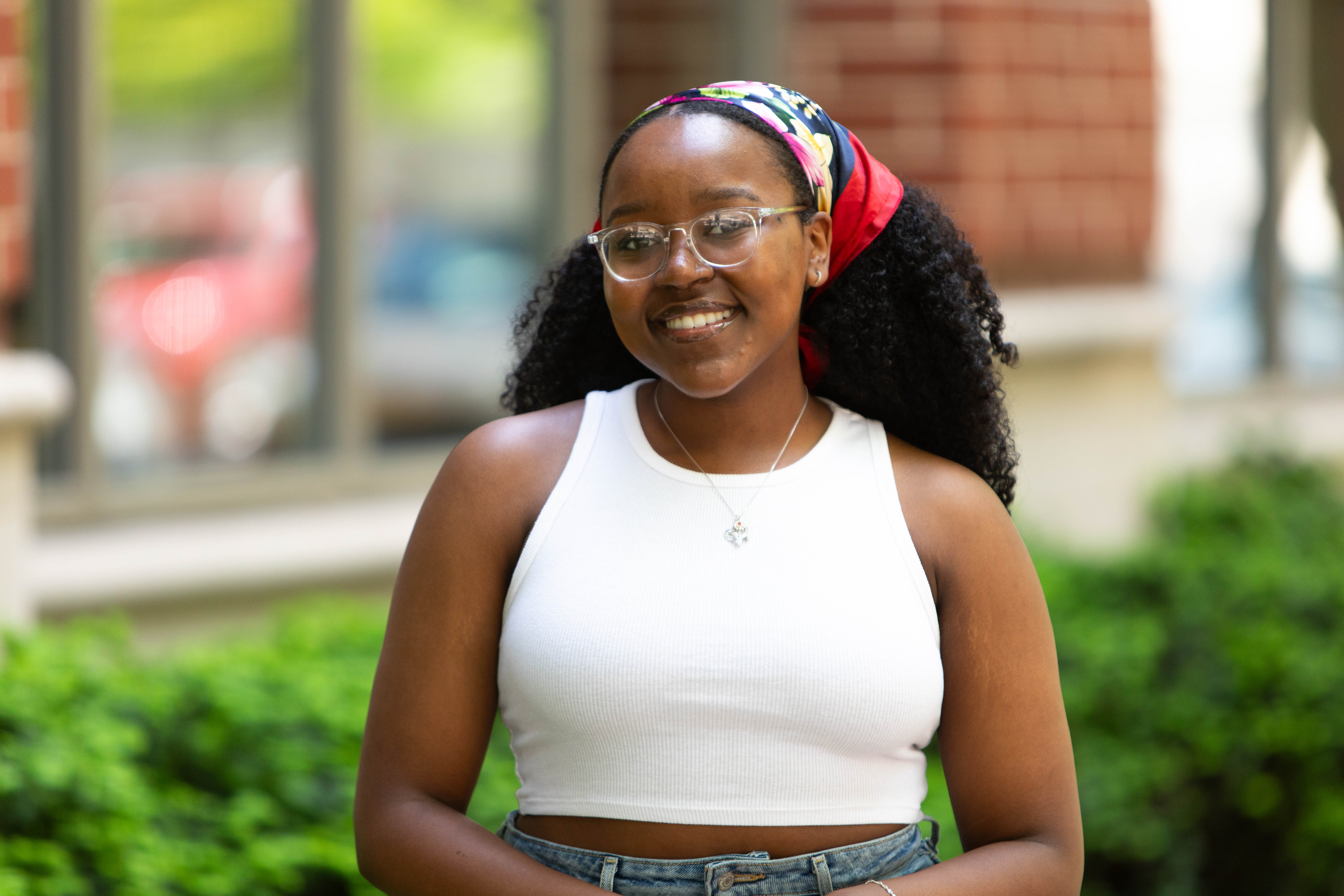 An image of Mya Wraggs with a white shirt and colorful hair scarf standing outside with a blurred background.