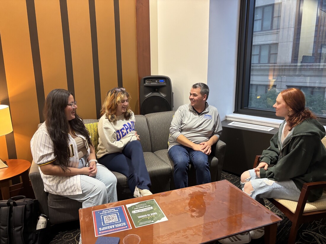 A group of people sit around a coffee table in a cozy lounge area; a city building is visible out the window