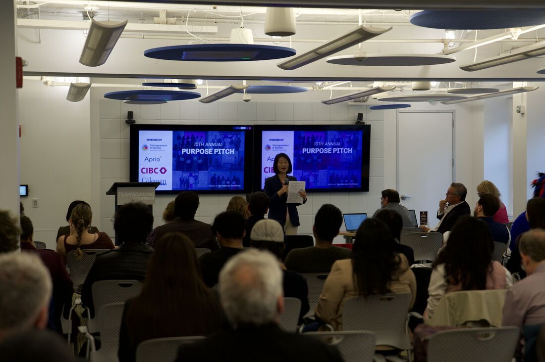 at the front of a packed conference room, a woman speaks in front of slides reading purpose pitch
