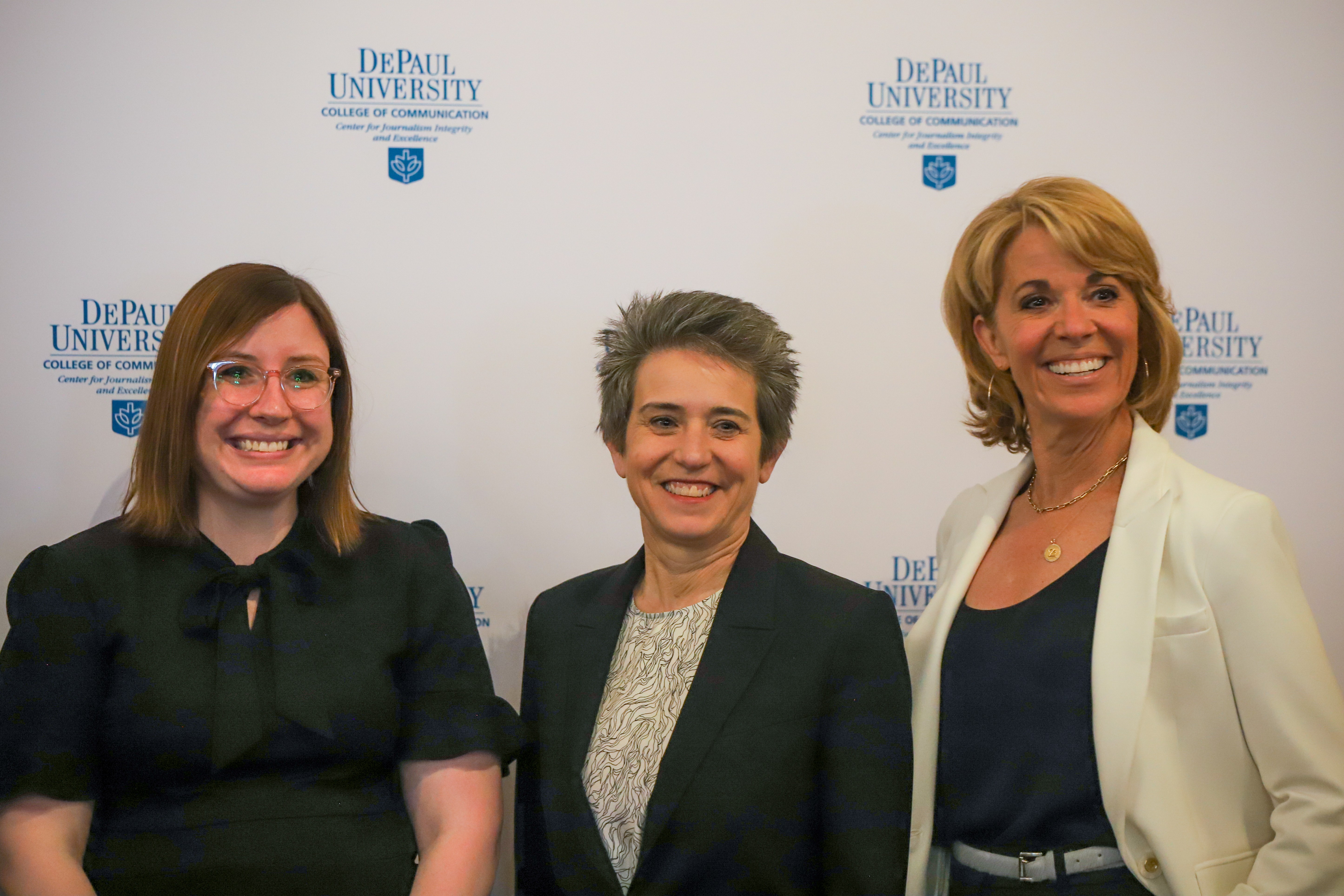 Three women in business attire smile standing in front of DePaul University signage.