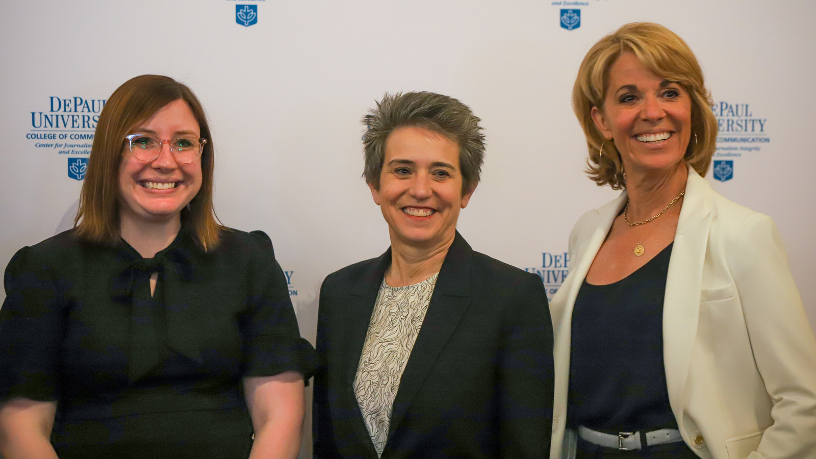 Three women in business attire smile standing in front of DePaul University signage.