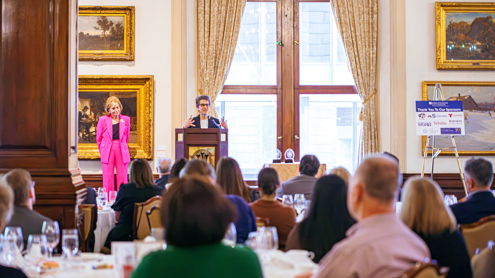 A woman stands at a podium and addresses a crowd seated at tables.