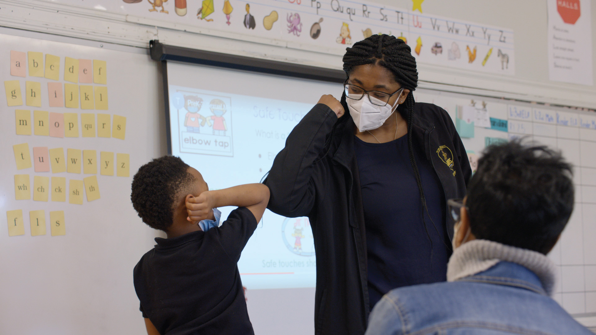 An elementary school teacher, Chelsea Bennett, who has braids, glasses, and a KN95 mask, elbow tapping a student wearing also wearing a mask.