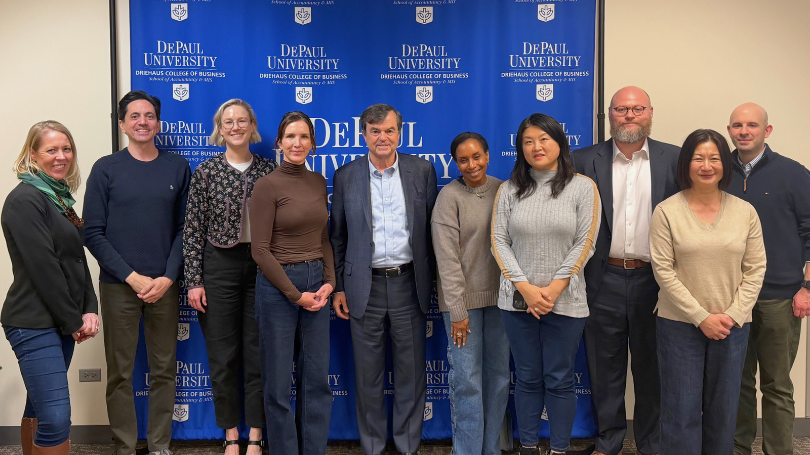 a group of DePaul faculty in dressy casual outfits pose with Jim Logothetis, a man in his sixties wearing a suit with a heart-shaped face and an affable grin; behind them, a bright blue banner displays the DePaul logo