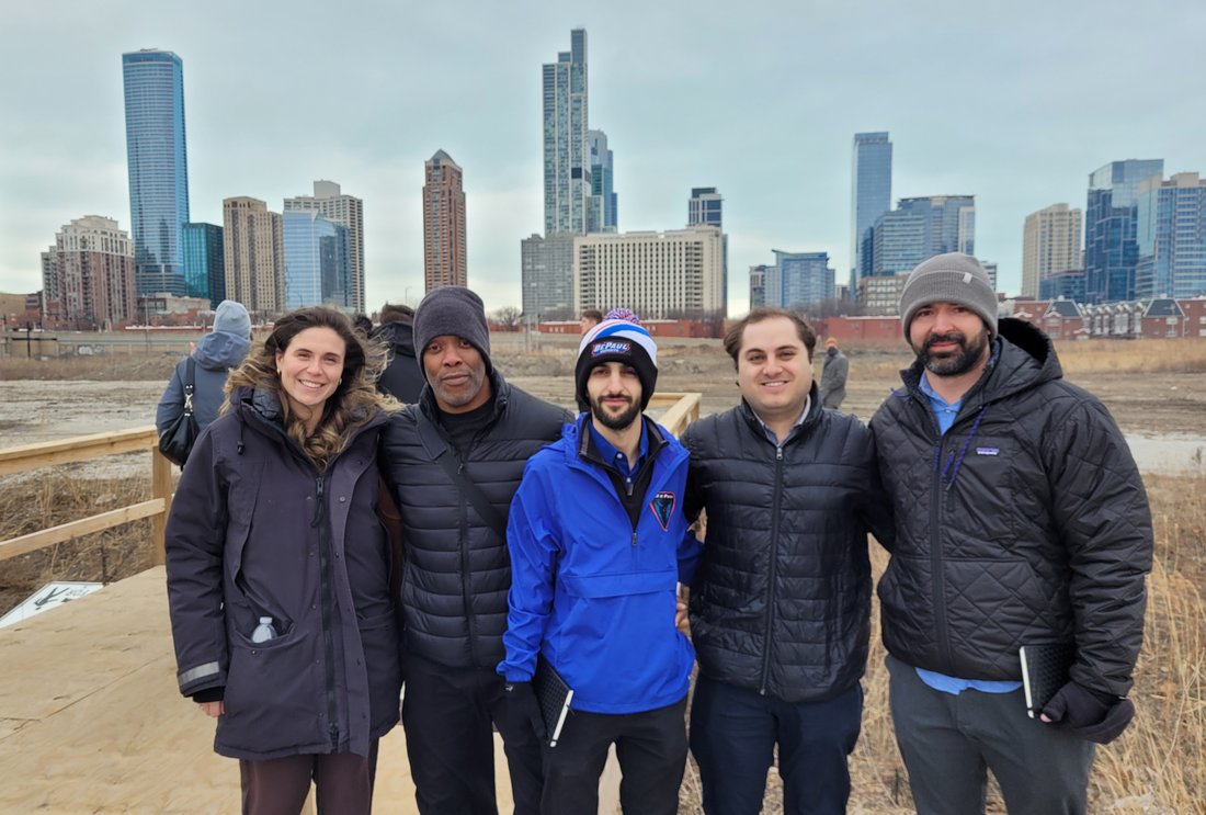 a group of five people in coats pose at an empty development site, with the skyline of Chicago's south loop in the background