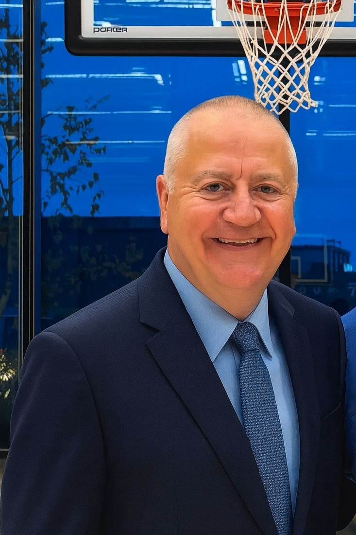 Scott Steffens, a white man in his sixties in a navy suit and tie, smiles warmly in front of a basketball hoop