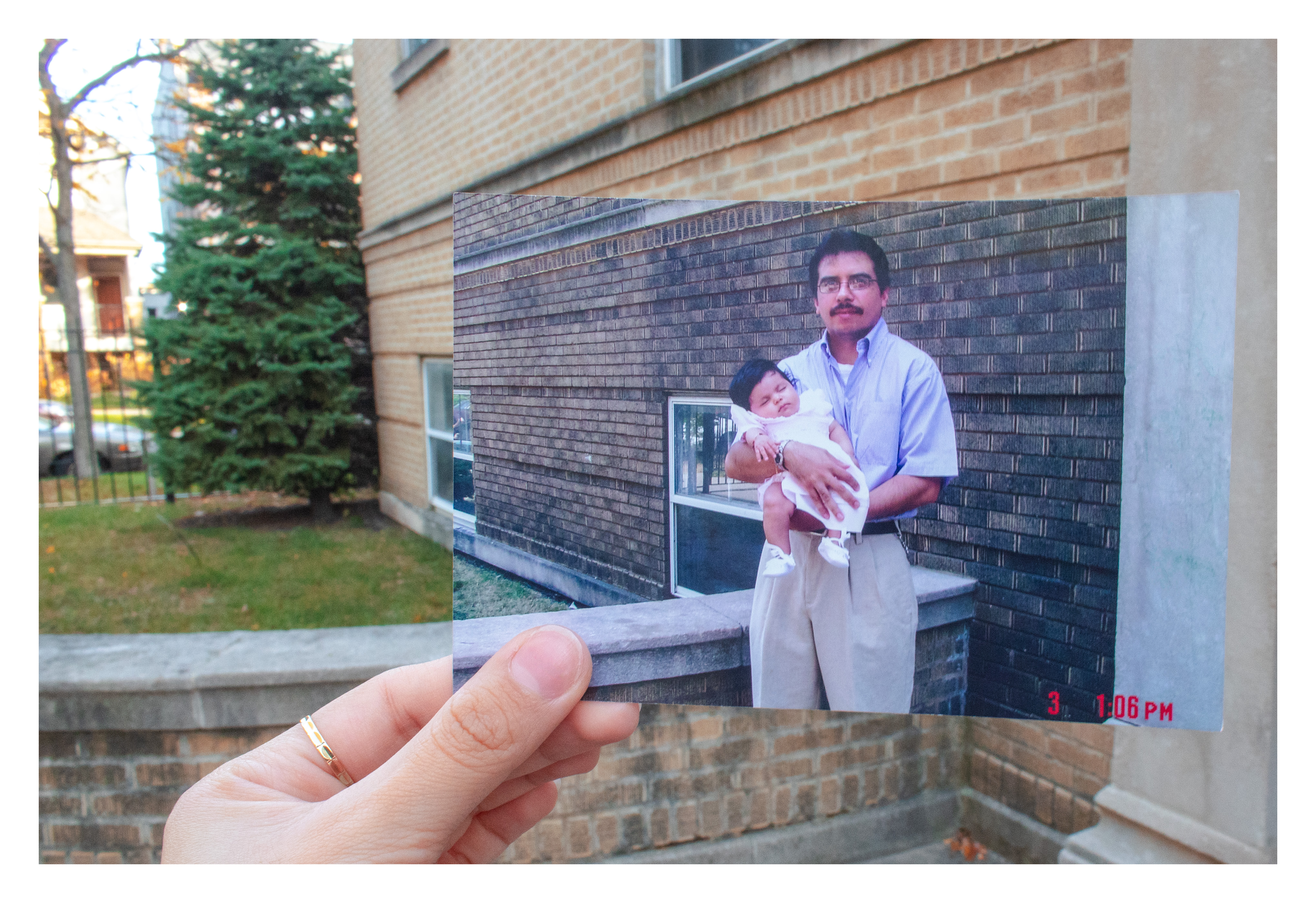 Daniella's hand holds up a childhood photo of her father holding her as a baby on a stoop. The building in the childhood photo lines up with the same building in the present day.