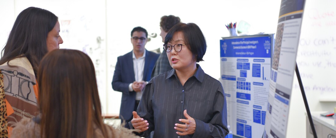 a woman in a collared shirt presents a research poster to onlookers, with other posters and presenters in the background