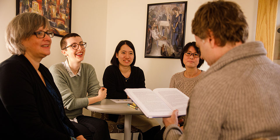 Four students listening to a faculty member while he reads from a book.