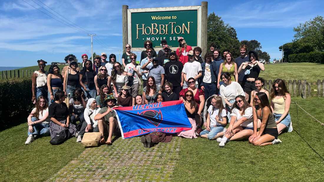 group of DePaul students posing in front of the Hobbiton movie set sign with a DePaul flag