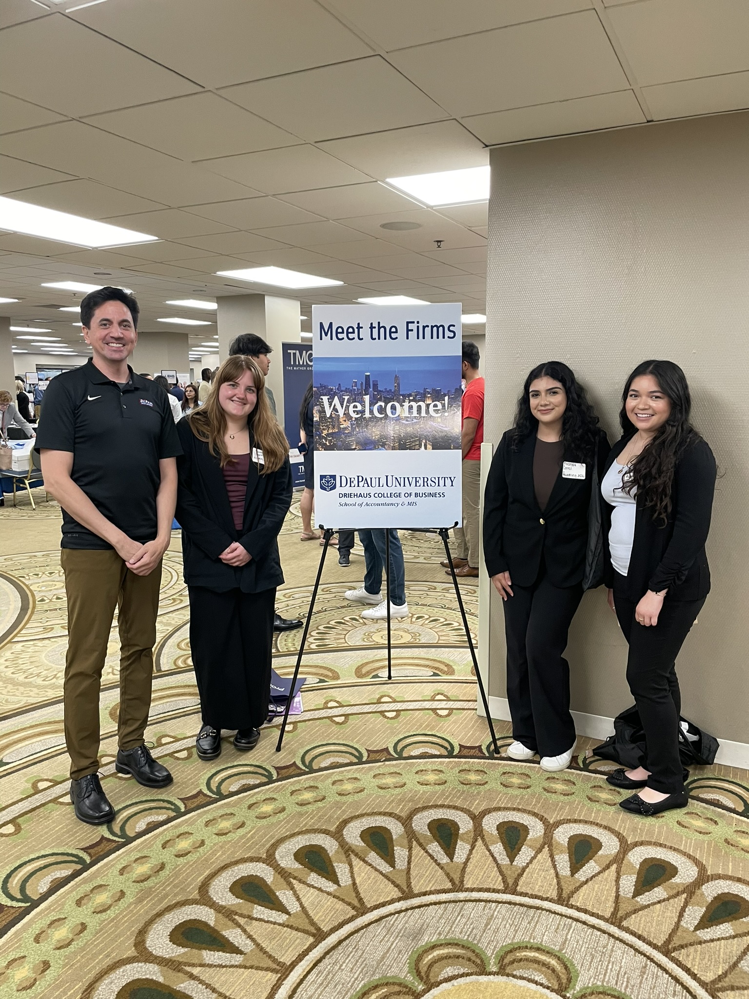 Three college students and a staff member pose in front of a sign that says "Meet the Firms." They are wearing business casual. The setting is a hotel conference center bustling with a job fair event.