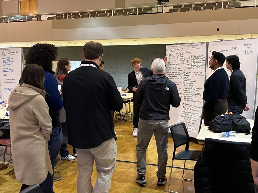 A crowd gathers around a student presenting a large, handwritten flow chart