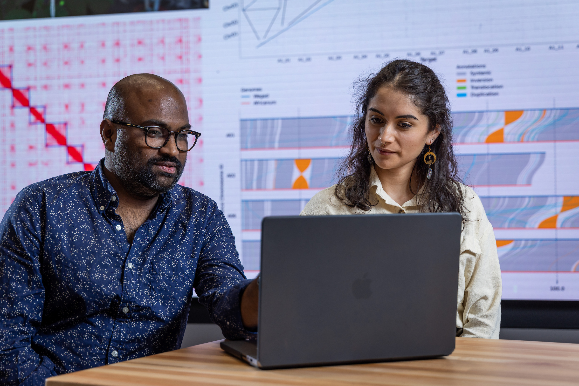 Two people in front of a laptop with a screen of graphs behind them