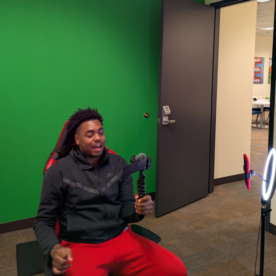 a young man sits in a chair filming himself with a phone on a stabilizing stand, plus a ring light