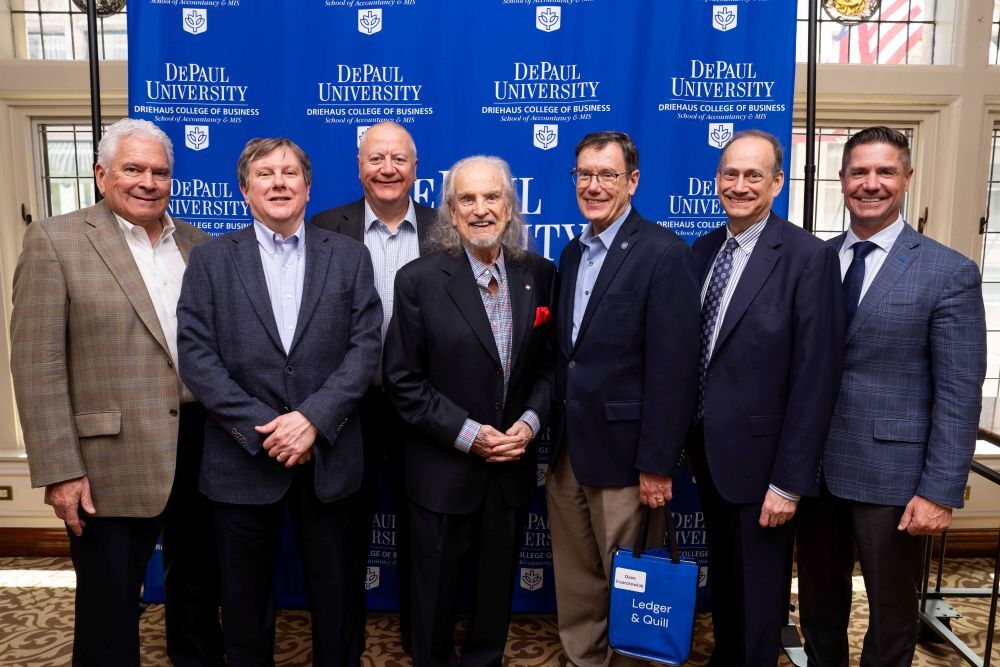 seven older white men in crisp suits smile in front of a vibrant blue depaul photo backdrop