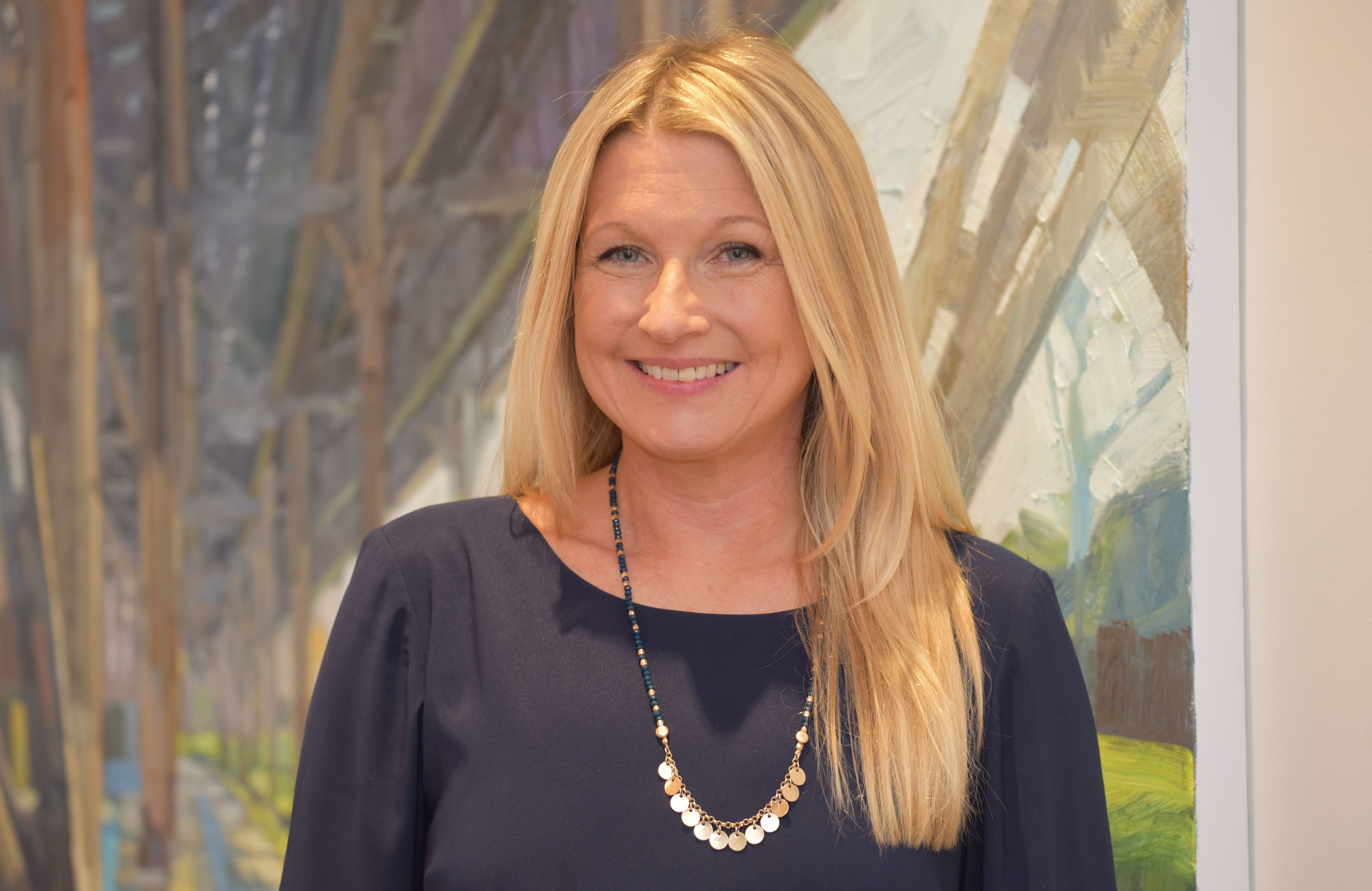 a white woman in her forties with long blond hair against a painted forest scene