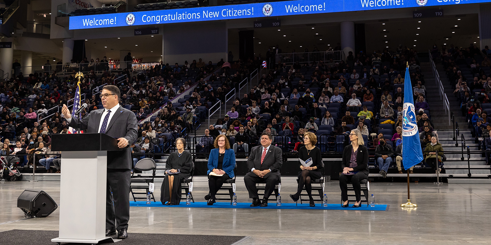 President Rob Manuel stands at a podium in the center of an arena floor, behind him sits dignitaries.