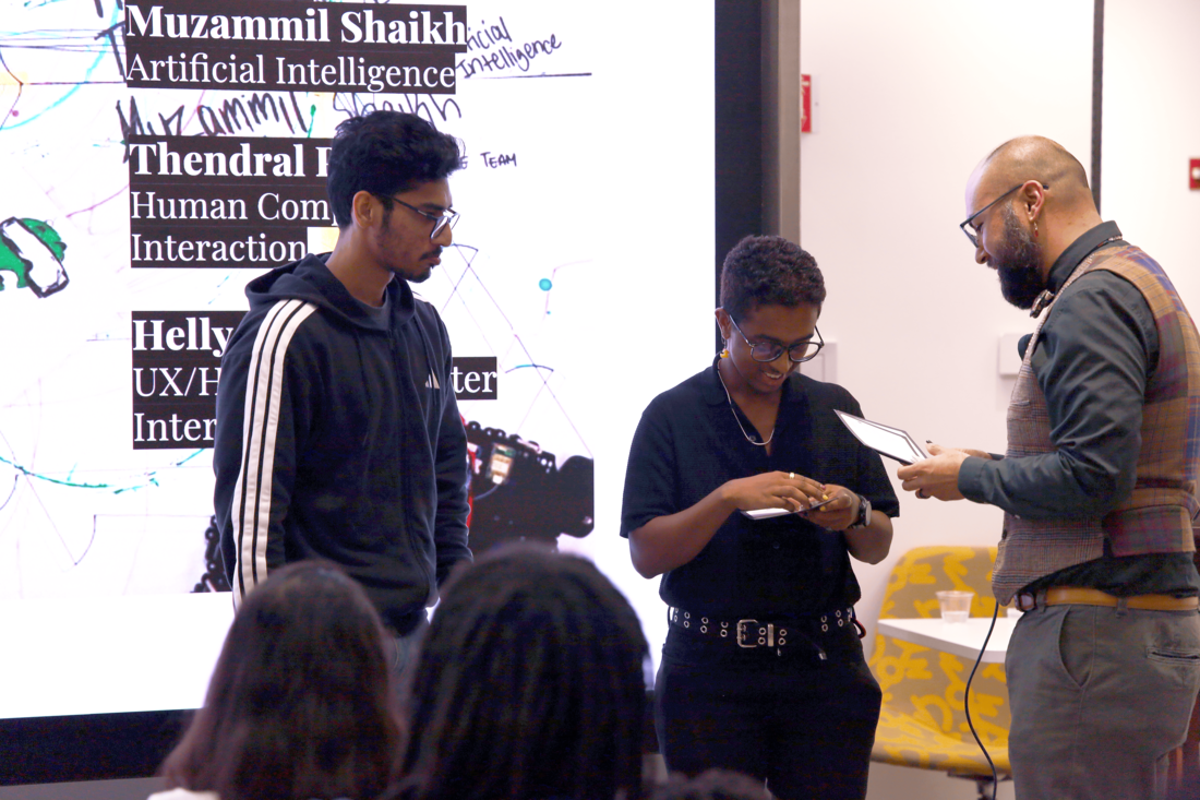 Two students on the left in front of a large screen receiving a coin passed from a faculty memer on the right