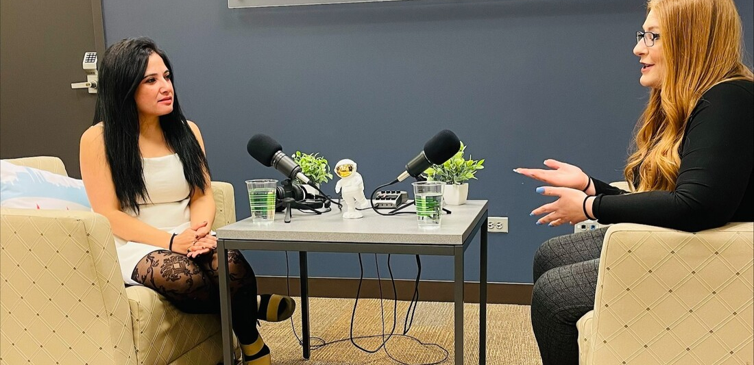 two podcasters chat at a table with large mics with a BETA hub sign on the wall behind them