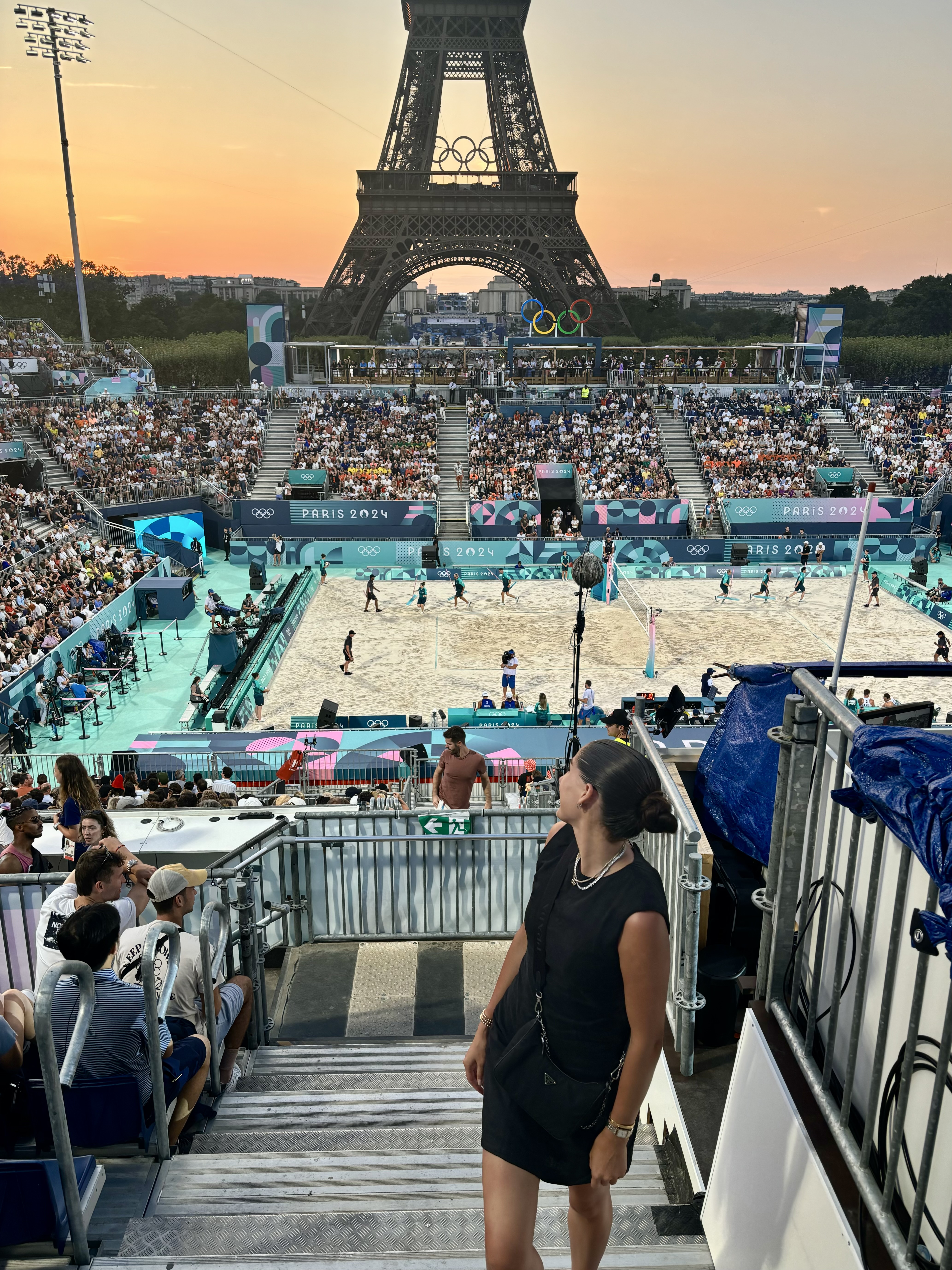 A beach volleyball court at sunset. The Eiffel tower looms in the background and, in the stands in the foreground, a young woman looks at it over her shoulder