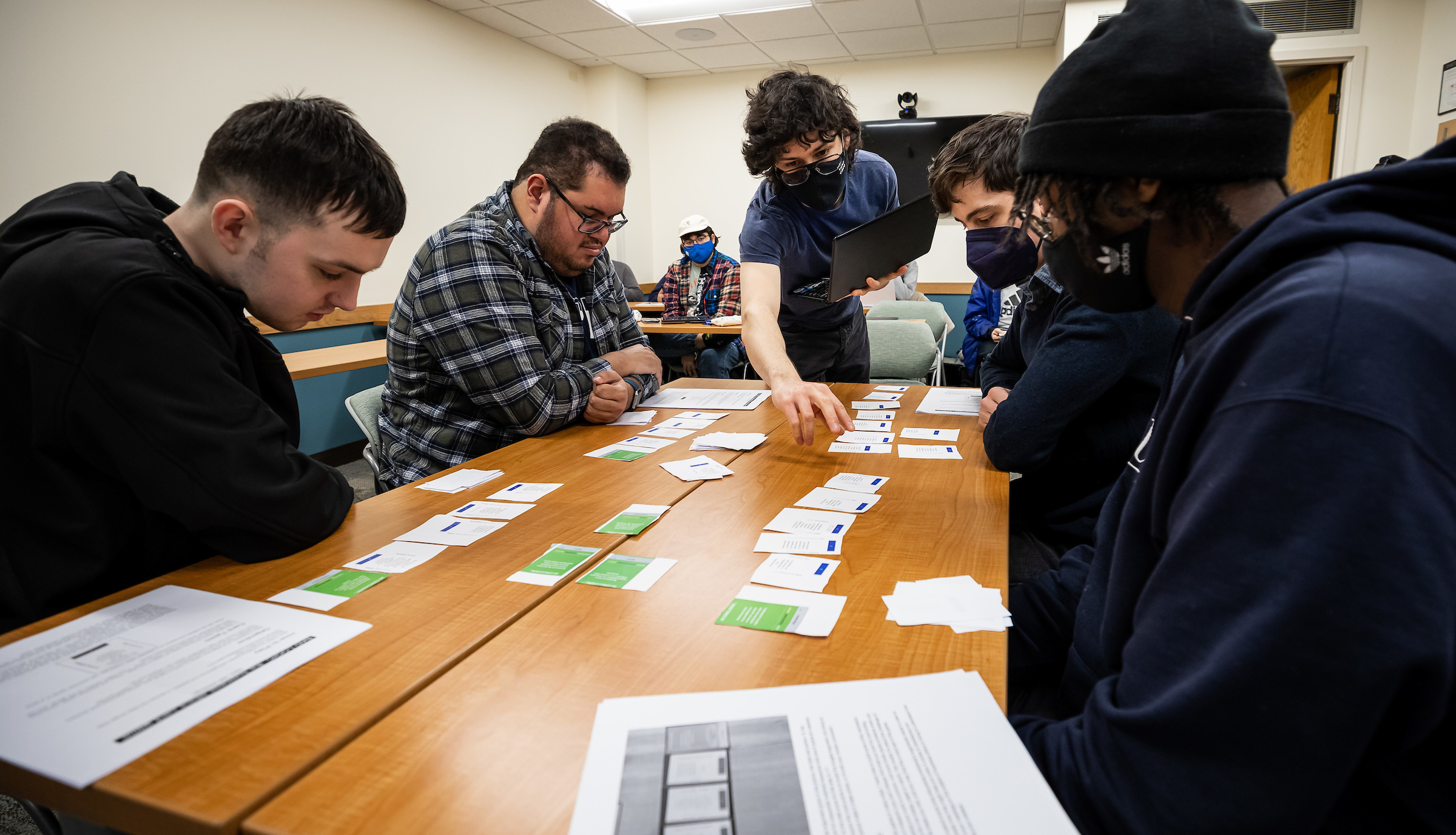 Students sit around a table looking at cards lined up for a game. One student holds a laptop.