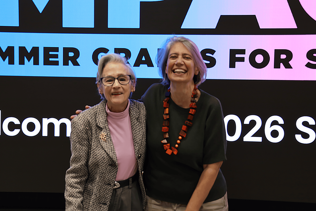 Two white women smile and pose for the camera in front of a large LED screen