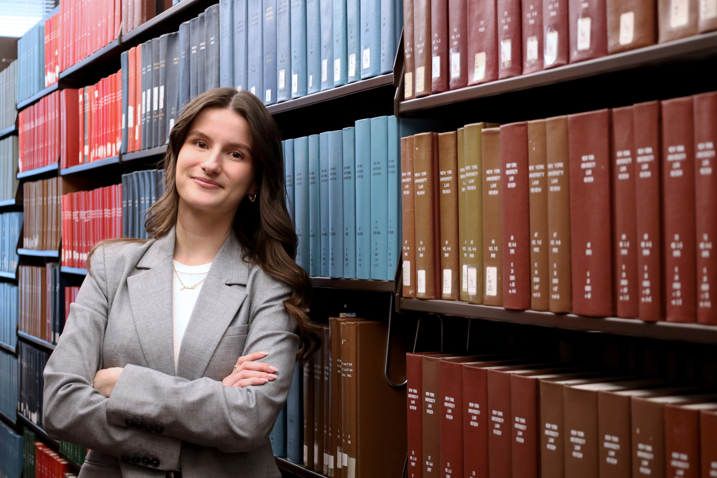Smiling Law Student, Amna, standing next to Library book case with arms crossed in a proud stance