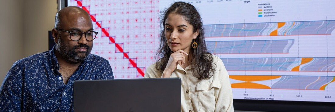 faculty and student working on laptop with large screen visualizing data behind them