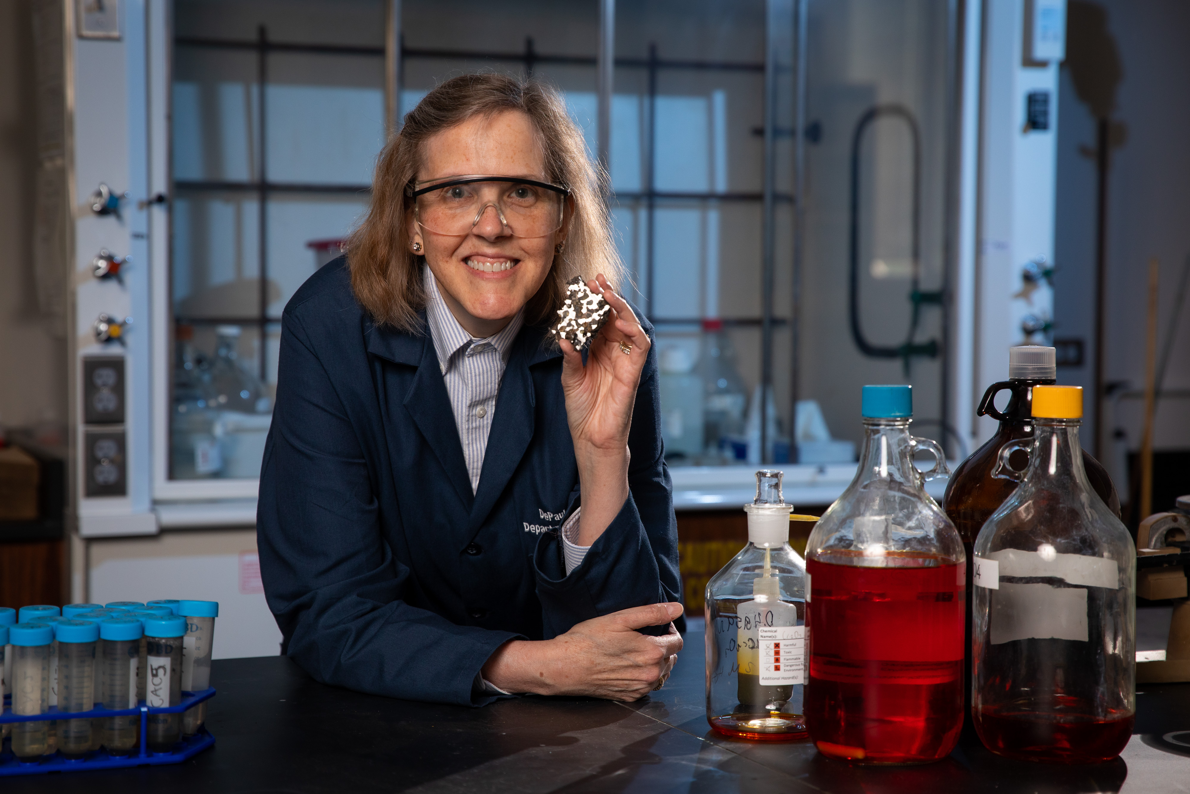 Wendy Wolbach wears safety goggles and a blue lab coat, holding a small shiny meteorite and surrounded by glass containers with chemicals in her lab.