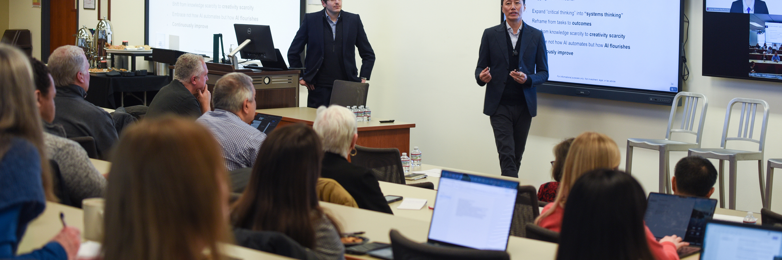 Speakers in suits pace in the front of a classroom packed with middle aged professionals