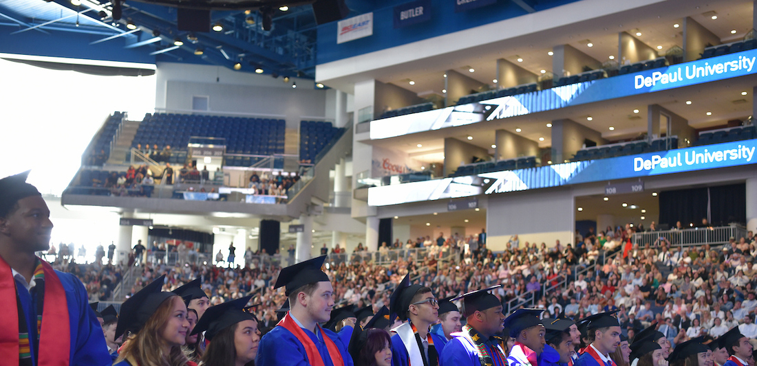 a line of students in graduation caps and vibrant blue gowns look hopefully ahead, with a packed stadium behind them