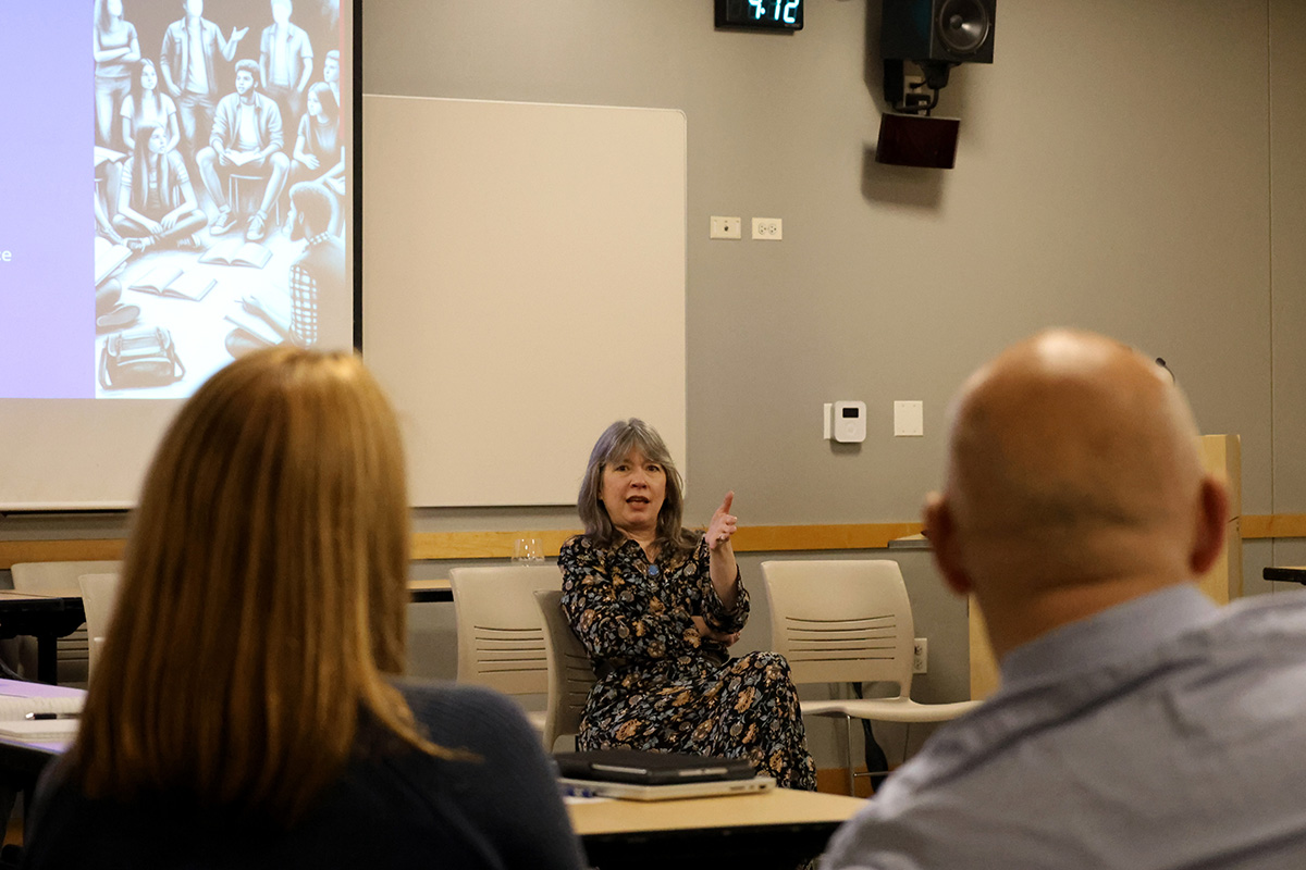 A faculty member sits in the front of a room talking, while others look on.