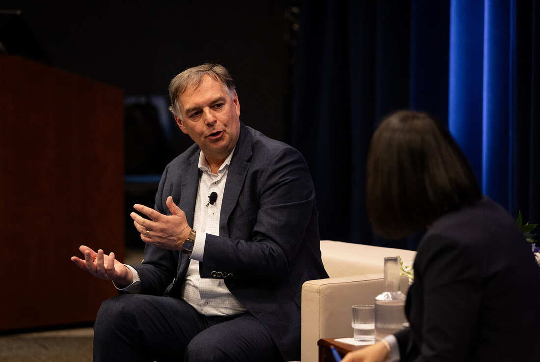 a white man in his early sixties in a suit with a lapel mic gestures as he speaks on a dramatically lit stages, sitting opposite another person in an armchair