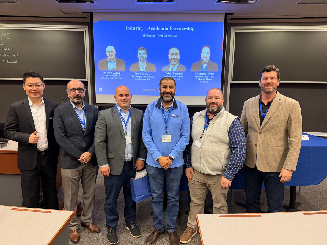five people in business casual wearing lanyards pose at the front of a classroom