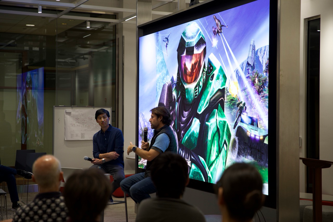 Two men in conversation in front of a video wall displaying a video game