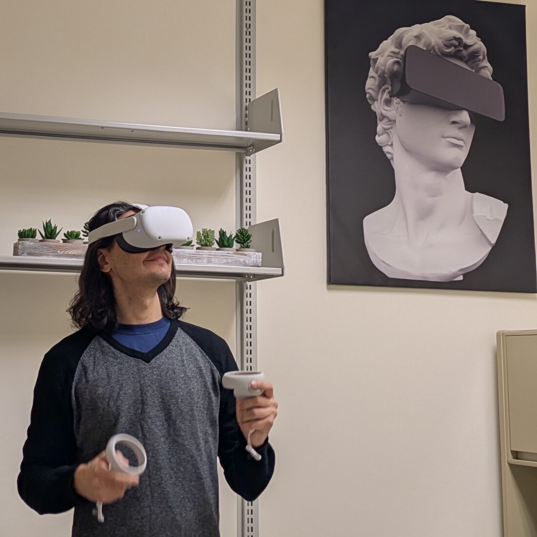 a man in a VR headset holding two controllers looks off in the distance; behind him on the wall, there's a graphic of a roman bust wearing a similar headset 