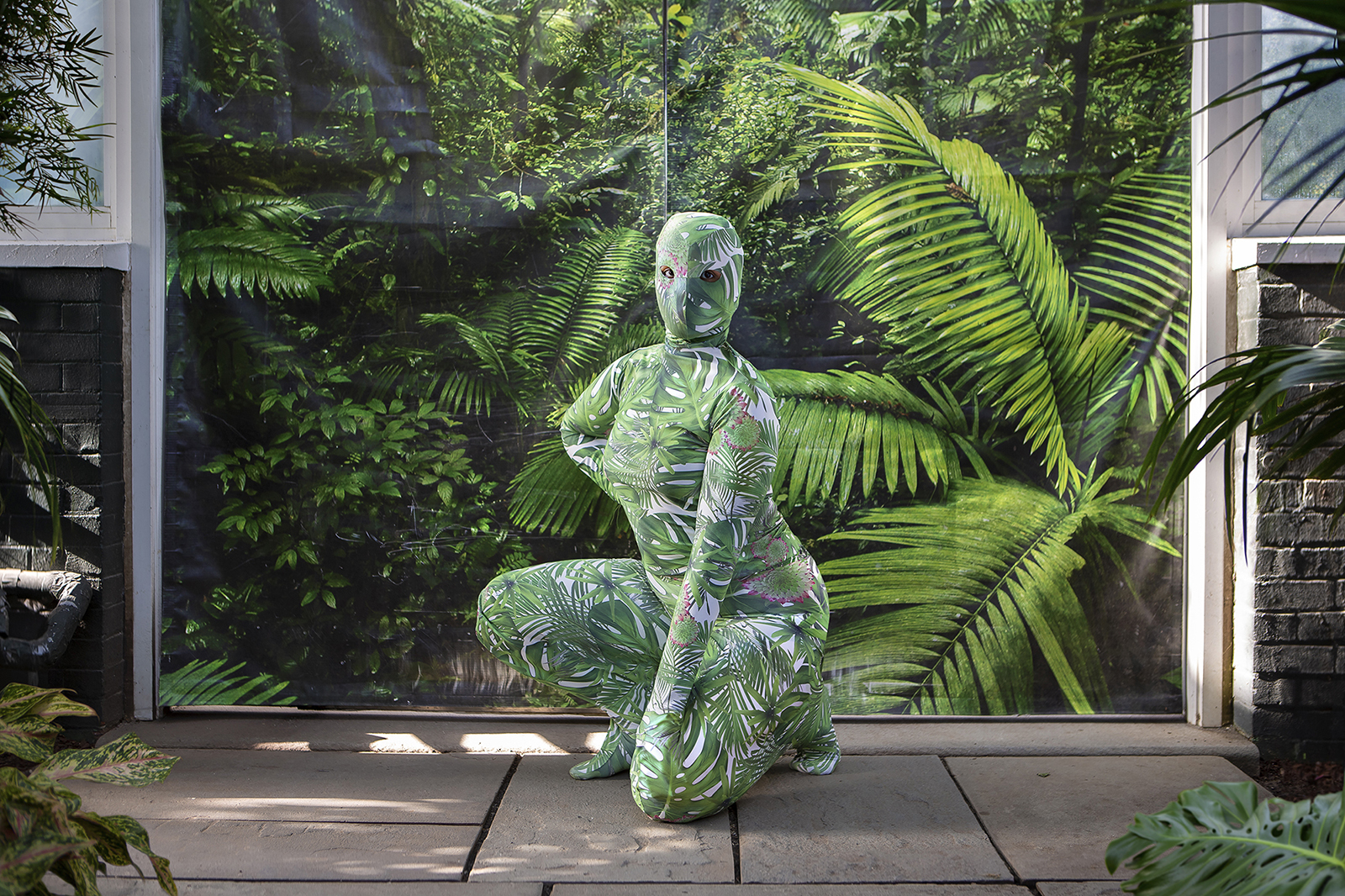 A figure in a suit covered in leaf print kneels in front of a window with leaves behind it.