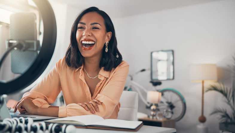 Social media influencer sitting at a table in front of a phone with a ring light, laughing and talking to her online followers