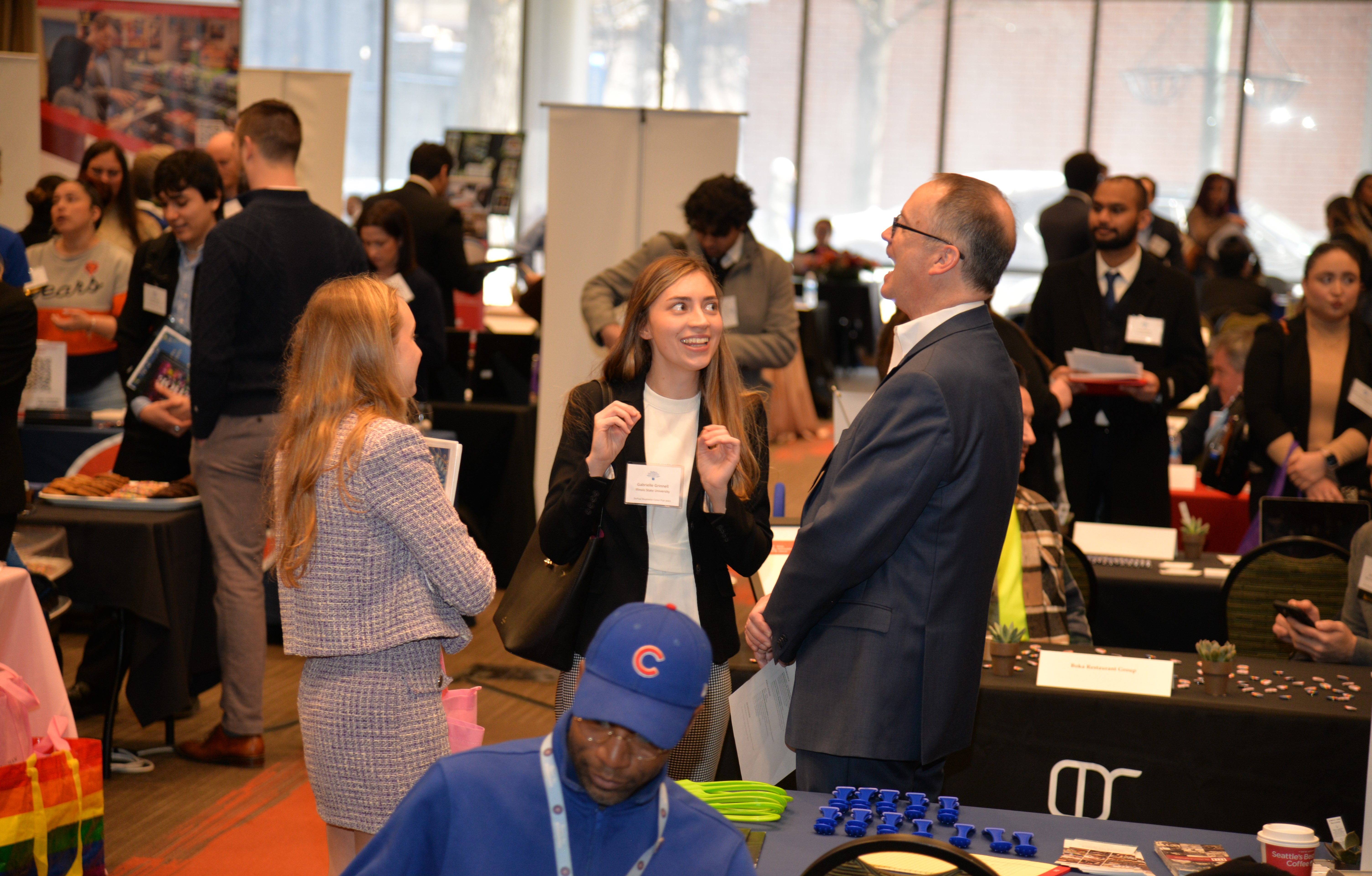 A group of students and professionals in business casual laugh in the middle of a hall crowded with employers' tables and networking students