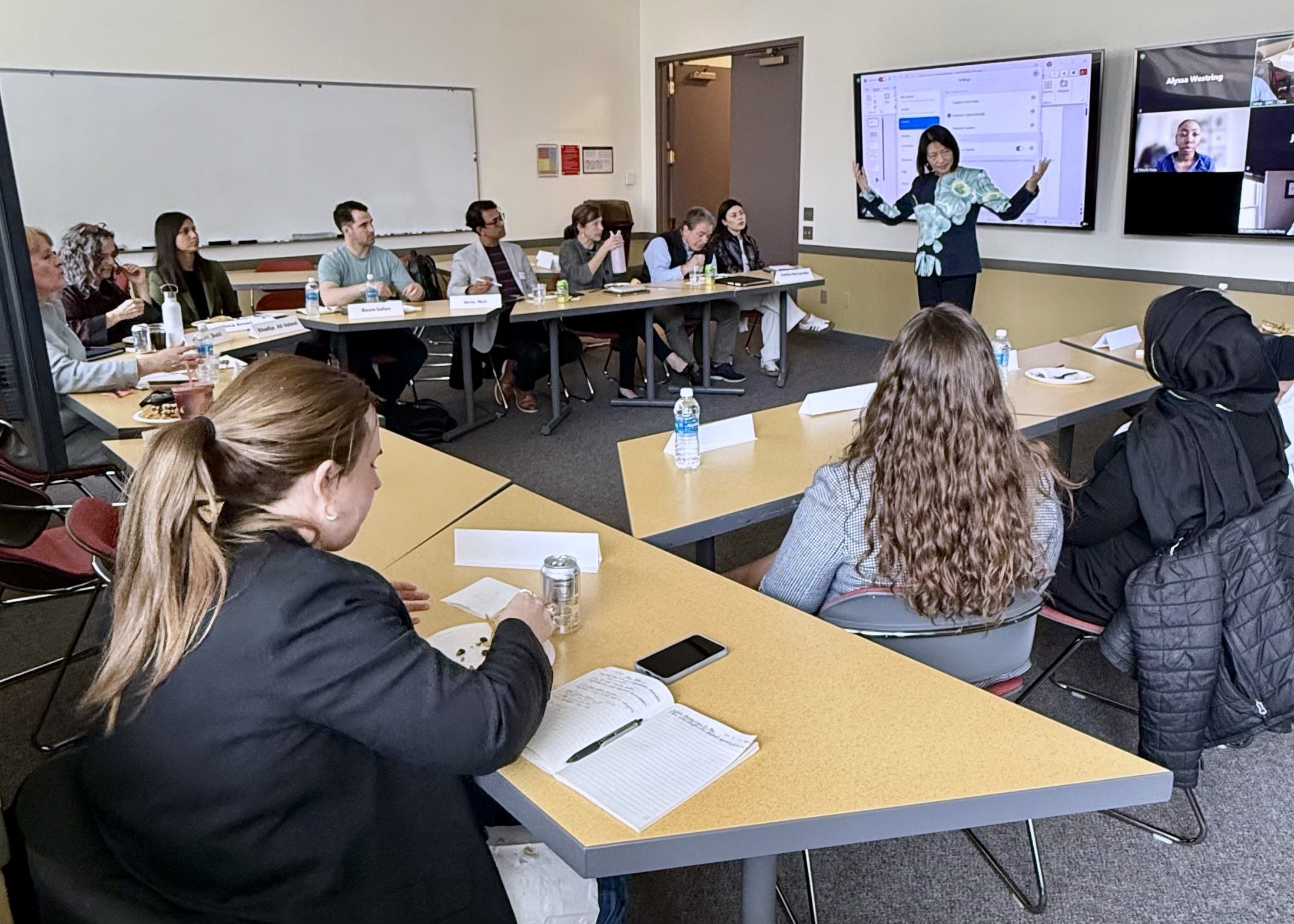 A group of professionals eats lunch at a u-shaped arrangement of tables as they watch a presentation and others join in on Zoom