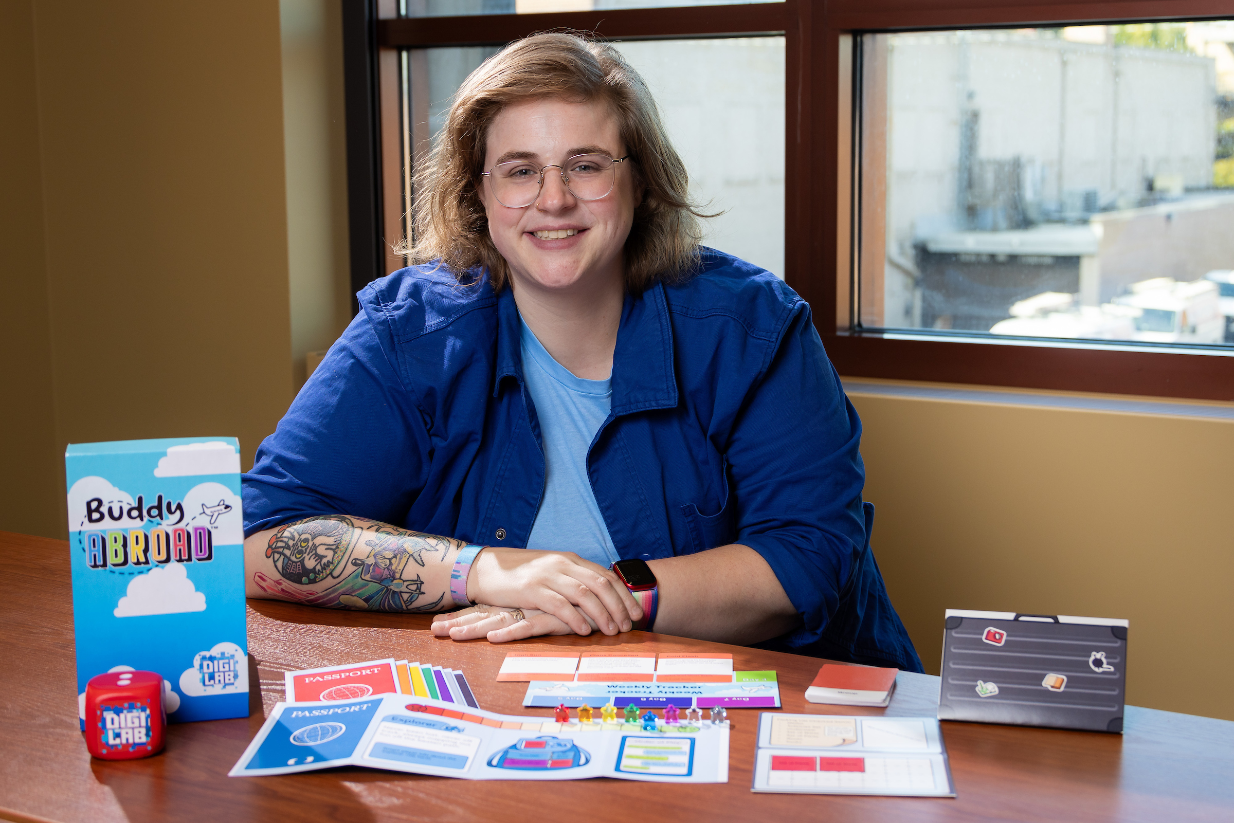 Person with blond hair and glasses smiles while holding a game piece and has a colorful board game in front of her with the name Buddy Abroad on the box