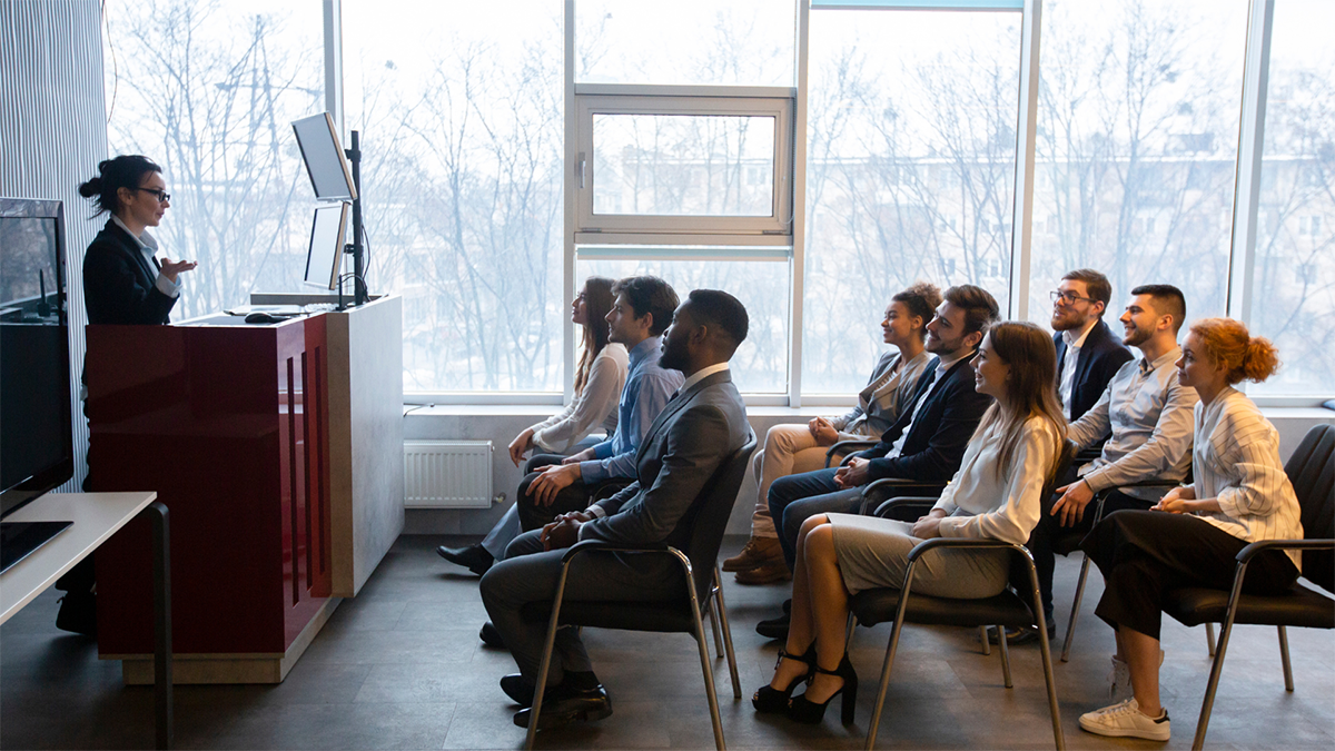 An image of a woman standing behind a podium talking to a small group of people sitting in chairs.